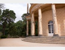 Detailaufnahme eines historischen Gebäudes mit säulengetragenem Portikus, großen Rundbogenfenstern und einer Treppenanlage vor dem Eingang. Im Hintergrund befinden sich Bäume und ein geschotterter Vorplatz.