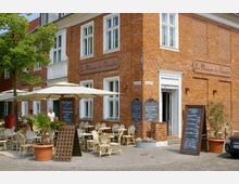 Rotes Backsteingebäude mit dem Schriftzug „La Maison du Chocolat“, davor ein Cafébereich mit Korbstühlen, Tischen und Sonnenschirmen. Mehrere Tafeln mit handgeschriebenen Menüpunkten stehen vor dem Eingang und auf dem gepflasterten Platz.
