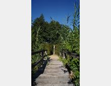 Holzbrücke, die über eine von Pflanzen umgebene Fläche führt, mit hohen Gräsern und Büschen am Rand, die zu einer sandigen Fläche und einer Statue im Hintergrund führt; im Hintergrund dichter Baumwald und klarer blauer Himmel.