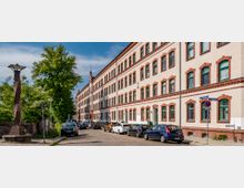 Straßenansicht eines historischen Gebäudes mit vier Etagen, verputzter Fassade und dekorativen roten Klinkerrahmen um die grünen Fenster. Vor dem Gebäude stehen geparkte Autos, ein Gehweg mit Bäumen und eine Backsteinlaterne sind ebenfalls sichtbar.