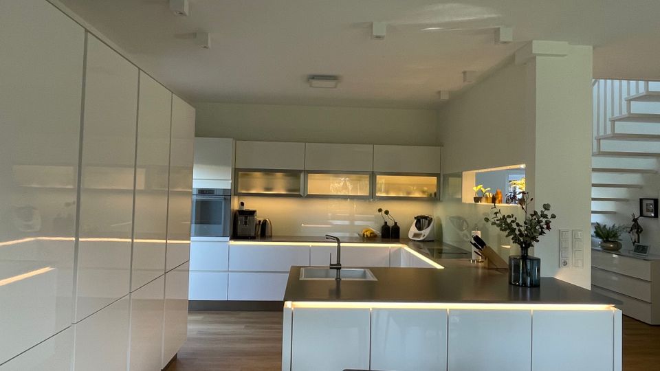 Moderne Küche mit glänzenden, weißen Einbauschränken und einer zentralen Kücheninsel mit grauer Arbeitsplatte und integriertem Waschbecken. Helle LED-Beleuchtung unter den Schränken und der Insel sorgt für indirektes Licht, während eine offene Treppe und Dekorationsgegenstände im Hintergrund sichtbar sind.