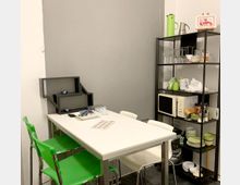 Ein kleiner Gemeinschaftsraum mit einem weißen Tisch, umgeben von grünen und weißen Stühlen. Im Hintergrund steht ein schwarzes Regal mit Geschirr, Gläsern, einer Mikrowelle und Küchenutensilien.