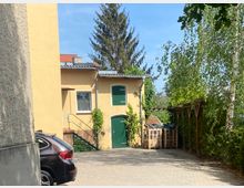 Pflasterhof mit einem gelben Gebäude, das grüne Türen und Fensterläden hat. Links ist ein geparktes Auto, rechts ein überdachter Bereich neben Bäumen und Sträuchern, im Hintergrund große Nadelbäume.