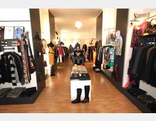 Ein moderner Bekleidungsladen mit Holzfußboden, Regalen und Kleiderstangen an den Wänden sowie einem zentralen Tisch mit gefalteter Kleidung. Die Beleuchtung besteht aus runden Hängelampen, und die Ware umfasst verschiedene Kleidungsstücke, Accessoires und Schuhe.