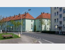 Straßenecke in einem Wohngebiet mit mehrstöckigen, pastellgrünen Wohnhäusern mit roten Ziegeldächern, umgeben von Bäumen und Gehsteigen. Auf dem Schild ist der Straßenname "Große Hagenstraße" zu sehen, und auf dem Grünstreifen stehen Bänke.