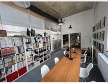 Ein heller Arbeitsraum mit langen Holztischen, weißen Stühlen und mehreren schwarzen Schreibtischlampen. An der linken Wand befinden sich offene Regale mit Aktenordnern und Büchern, während die rechte Wand mit gerahmten Bildern dekoriert ist; im Hintergrund sind Glaswände und ein angrenzender Bereich sichtbar.