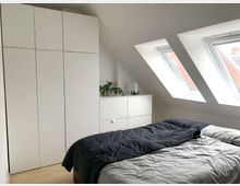 Helles Schlafzimmer mit Dachschrägen, zwei Dachfenstern, einem Doppelbett mit dunkelgrauer Bettdecke, einem weißen Kleiderschrank und einer weißen Kommode, auf der eine Pflanze und eine Lampe stehen. Der Boden besteht aus hellen Holzplanken.