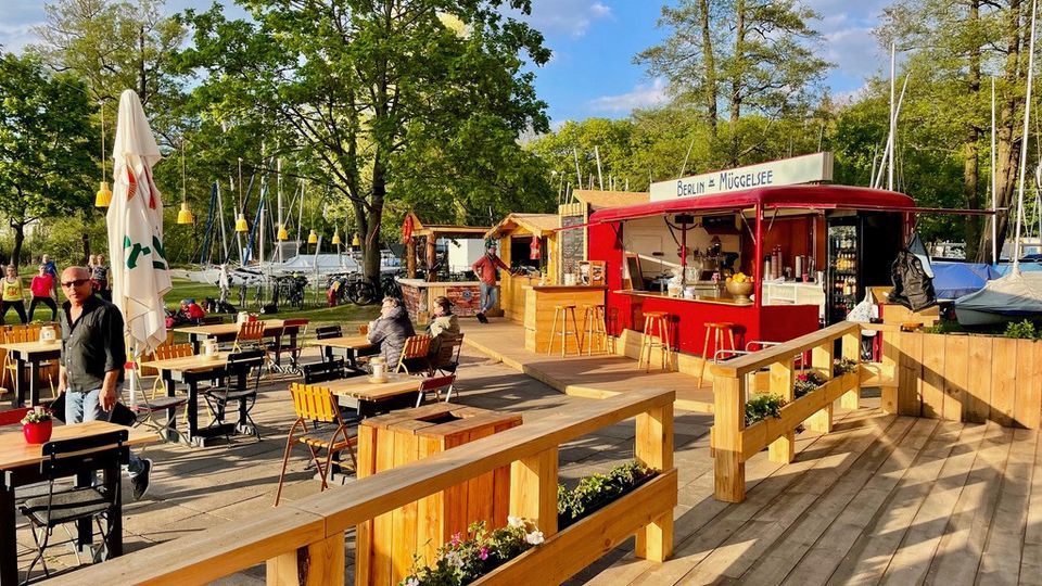 Ein Outdoor-Cafébereich mit Holzmöbeln und einer roten Imbissbude, beschriftet mit "Berlin Müggelsee", umgeben von großen Bäumen und einem nahegelegenen Segelbootanleger. Im Vordergrund befinden sich Pflanzenkästen und sonnendurchflutete Terrassenplatten.