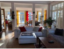 Helles Wohnzimmer mit weißen Sofas, Holzfußboden und großen Fenstern, die mit gelben und roten Vorhängen dekoriert sind. Im Hintergrund befindet sich ein Essbereich mit einem Holztisch und Stühlen sowie eine offene Küche mit roten Schränken.