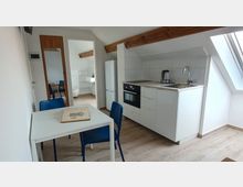 Heller Dachgeschossraum mit kleiner Küchenzeile in Weiß, integriertem Backofen, Kühlschrank und Spüle unter einer Dachschräge mit Dachfenster. Im Vordergrund steht ein weißer Esstisch mit blauen Stühlen; sichtbare Holzbalken und eine Türöffnung führen in ein angrenzendes Bad.