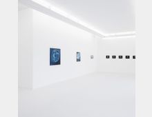 Ein minimalistischer Galerieraum mit weißen Wänden und einem glatten, hellen Boden. Kunstwerke, darunter Gemälde und kleine Bilder, sind in einem geraden Abstand an den Wänden angebracht; die Beleuchtung erfolgt über lange, schlichte Deckenleuchten.