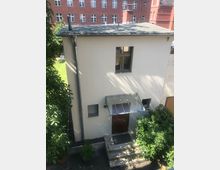 Kleines, zweigeschossiges Gebäude mit heller Fassade und Flachdach, mit einem zentralen Fenster im Obergeschoss und einem Eingang im Erdgeschoss unter einem gläsernen Vordach. Vor dem Eingang führen wenige Stufen hinauf, umgeben von Büschen und Bäumen; im Hintergrund ist ein größeres rotes Backsteingebäude mit vielen Fenstern zu sehen.