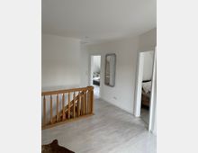 Wohnbereich in einem Obergeschoss mit hellem Laminatboden, einer Holztreppe mit Geländer, zwei offenen Türen zu angrenzenden Zimmern und einem rechteckigen, silbernen Wandspiegel. Die Wände sind in Weiß gehalten.