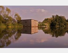 Holzhütte am Ufer eines ruhigen Sees, umgeben von Bäumen und Schilf, mit einer klaren Spiegelung der Szenerie im Wasser unter einem leicht bewölkten Himmel.