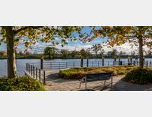 Sitzbank an einer Uferpromenade mit Blick auf einen ruhigen Fluss, umgeben von Bäumen mit herbstlich gefärbtem Laub. Im Hintergrund sind ein Steg mit Geländern und eine grüne Landschaft zu sehen.