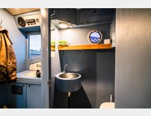 Kompakter Innenraum eines Schiffskajüte-ähnlichen Raumes mit blau gestrichenen Wänden, einem runden Bullauge, einem kleinen Edelstahlwaschbecken und einer hölzernen Ablage. An der Wand hängt eine braune Lederjacke, und auf der Arbeitsfläche befinden sich Karten und ein Fernglas.