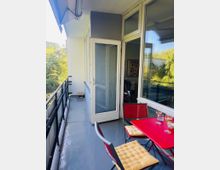 Schmaler Balkon mit grauem Boden und schwarzem Metallgeländer, angrenzend an eine Wohnung mit großen Fenstern und einer geöffneten Glastür. Rechts steht ein roter Tisch mit Stühlen und Sitzkissen; im Hintergrund ist eine grüne Baumlandschaft zu sehen.