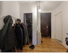 Ein schmaler Flur mit Holzboden und weißen Wänden. Links steht ein Kleiderständer mit hängenden Jacken, vorne eine dunkle Holzeingangstür, rechts ein kleiner Abstellraum mit Regalen.