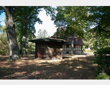 Holzgebäude mit flachem Dach im Vordergrund, umgeben von hohen Bäumen und trockenem Waldboden; im Hintergrund ein Fachwerkhaus mit Ziegeldach in einer parkähnlichen Umgebung.