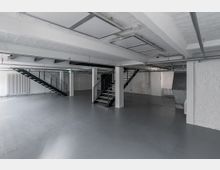 Industrie-ähnlicher Innenraum mit einer offenen Fläche, grauem Boden und weißen Wänden aus Ziegeln. Zwei schwarze Metalltreppen führen zu einer oberen Ebene, und an der Decke sind freiliegende Leitungen sichtbar.