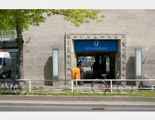Eingang zur U-Bahn-Station "Nollendorfplatz" in Berlin, mit einem blauen Schild über dem Durchgang. Der Eingang ist von einer grauen Betonwand umgeben, die mit ornamentalen Verzierungen eingerahmt ist, und im Vordergrund stehen Fahrräder sowie Menschen neben einem Fußweg.