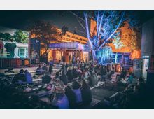 Ein Open-Air-Kino bei Nacht mit einer großen Leinwand links, auf der ein Film mit Untertiteln läuft. Im Vordergrund sitzen Zuschauer auf Bänken, umgeben von Bäumen und einer beleuchteten Holzhütte im Hintergrund.
