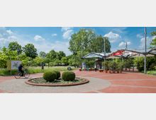 Eingangsbereich eines Parks mit gepflasterten Wegen, einem bepflanzten Rondell und einem überdachten Torbogen. Im Hintergrund sind Bäume und Wiesen zu sehen, während Besucher zu Fuß oder mit dem Fahrrad unterwegs sind.