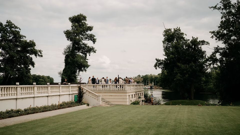 Weiße steinerne Terrasse mit Balustrade und Treppe in einem Park, davor eine große Rasenfläche. Rechts verläuft ein Fluss entlang dichter Bäume, im Hintergrund sind weitere Grünflächen unter bewölktem Himmel zu sehen.
