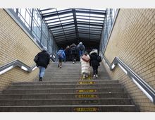 Eine überdachte Treppe in einem öffentlichen Durchgang mit gelben Backsteinwänden und Metallgeländern, auf der mehrere Personen nach oben gehen. Der Treppenbereich ist teilweise beschriftet, und das Dach besteht aus Metallträgern und Glas.