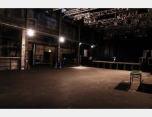 Eine leere Veranstaltungshalle mit industriellem Charakter, sichtbar durch freiliegende Metallträger, eine hohe Decke mit Beleuchtungseinrichtungen und schwarze Vorhänge im Hintergrund. Im Raum befindet sich eine Bühne, Lautsprecher und vereinzelte Tische.