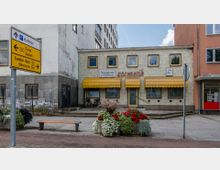 Fassade eines kleinen Gebäudes mit gelben Markisen, gelb-blauen Fliesen und der Beschriftung „Kosmetik“. Im Vordergrund befindet sich ein bepflanzter Blumenbeetkasten mit bunten Blumen sowie ein Straßenschild mit Wegweisern, daneben eine Sitzbank.