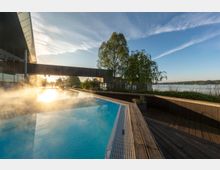 Außenbereich eines modernen Wellness-Spas mit einem dampfenden Infinity-Pool, umgeben von Holzterrassen, Bäumen und Blick auf einen ruhigen See im Hintergrund. Die tiefstehende Sonne erzeugt Reflexionen auf der Wasseroberfläche und beleuchtet die Umgebung.