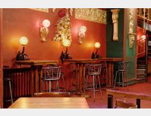 Raum im Stil eines nostalgischen Cafés mit roten Wänden, verziert mit goldenen, barocken Dekorationselementen. Hohe Holztische mit Metallhockern stehen vor Wandleuchten aus alten Telefonen und Kugellampen.