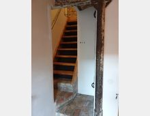 Treppenhaus mit einer schmalen Holztreppe, die nach oben führt, umgeben von weißen Wänden und einem freiliegenden Mauerwerkbereich mit Ziegelsteinen auf dem Boden und an der Wand. Ein rostiger Metallpfosten ist im Vordergrund sichtbar, und neben der Treppe befindet sich eine Tür mit der Markierung "WC2".