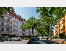 Straßenszene mit mehrstöckigen Wohnhäusern im typischen Stil des frühen 20. Jahrhunderts; links ein Gebäude mit weißen und roten horizontalen Streifen, rechts ein gelb-orangefarbenes Gebäude mit Balkonen. Die Straße ist mit geparkten Autos gesäumt und von hohen Bäumen überschattet.