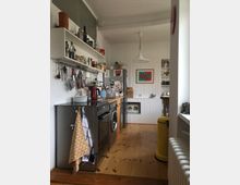 Kleine und schmale Küche mit Holzboden und weiß gestrichener Holzvertäfelung. Links befinden sich eine Arbeitsfläche mit Elektrogeräten und Regalen mit Küchenutensilien, während rechts ein gelber Mülleimer neben einer Heizung steht; im Hintergrund hängen Bilder an der Wand.