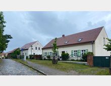 Straßenszene in einem kleinen Dorf mit Kopfsteinpflaster, gesäumt von Häusern mit roten Ziegeldächern und weißen Fassaden. Im Vordergrund ein Einfamilienhaus mit grünen Fensterläden und einem Vorgarten mit Zaun, umgeben von schmalen Gehwegen und Bäumen.