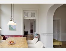 Wohnzimmer mit einem großen Esstisch aus Holz, zwei weißen Polsterstühlen und einer Schale mit Äpfeln. Im Hintergrund sind hohe weiße Doppeltüren und eine rote Couch sichtbar, umgeben von weiß gestrichenen Wänden und einer klassischen Raumaufteilung.