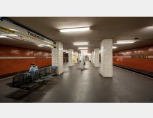 U-Bahn-Station "Yorckstraße" mit orangefarbenen Wandfliesen, weißen, gefliesten Säulen und dunklem Boden. Im Vordergrund befinden sich schwarze Sitzbänke, während im Hintergrund der Bahnsteig und Informationsschilder zu sehen sind.