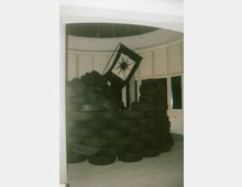 Ein Raum mit weiß gestrichenen Wänden und einer Reihe von Fenstern. Im Zentrum ist ein großer Stapel alter Autoreifen zu sehen, auf dem eine schwarze Flagge mit einem weißen Symbol hängt.