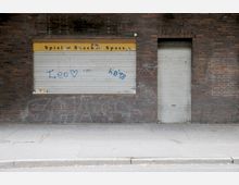 Backsteinfassade mit einem geschlossenen gelben Imbissfenster, auf dem "Spiel Snack Spaß" steht, und einer verschlossenen Metalltür rechts davon. Die Rollläden sind mit Graffiti bemalt, und der Gehweg davor ist leer.