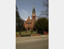 Backsteinkirche mit einem hohen, spitzen Turm und einer Uhr an der Front, umgeben von Bäumen. Vor der Kirche befindet sich ein eingezäunter Grünbereich mit einem dekorativen Tor aus Metall.