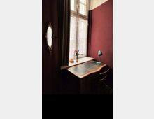 Ein kleiner Schreibtisch aus Holz mit lederner Schreibfläche steht vor einem hohen Fenster mit halbtransparenten Gardinen. Auf dem Fensterbrett stehen eine Vase mit Blumen und eine Lampe, die Wand ist dunkelrot gestrichen, und an der Seite hängt ein ovaler Wandspiegel.