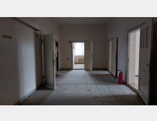 Ein leerer, ungepflegter Flur mit mehreren offenen Türen, die in kleine Räume führen. Der Boden ist mit grauen Fliesen bedeckt, und am rechten Rand steht ein roter Feuerlöscher. Im Hintergrund fällt Licht durch ein Fenster mit Gardine.