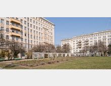 Mehrstöckige Wohngebäude im spätklassizistischen Stil, mit verzierten Fassaden und Balkonen, umgeben von einem gepflegten Park mit Rasenflächen, Blumenbeeten und Bänken, sowie einem säulenartigen Pavillon. Der Himmel ist klar und blau.