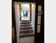 Blick durch einen schmalen, gemauerten Flur mit offenstehender Tür und aufsteigenden Ziegelstufen in einen Hof. Im Hof sind Pflastersteine, ein Sonnenschirm, Tisch und Stühle sowie ein Hund zu sehen; im Hintergrund befindet sich ein Gebäude mit großen Holztoren.