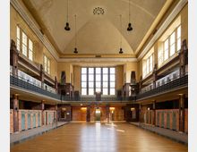 Historischer Innenraum eines ehemaligen Schwimmbades mit hoher Decke, großen Bogenfenstern und Geländern auf zwei Etagen. Die Wände sind mit Backsteinelementen und verzierten Umkleidekabinen im unteren Bereich gestaltet, während der Boden aus Holz besteht.