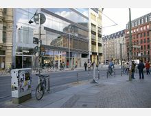 Straßenszene in einer urbanen Umgebung mit modernen Gebäuden aus Glas und Stahl sowie älteren Backsteinfassaden. Im Vordergrund sind Fahrräder, Fußgänger, öffentliche Verkehrsschilder und Kopfsteinpflaster zu sehen, während im Hintergrund Passanten und Straßenbahnschienen erkennbar sind.