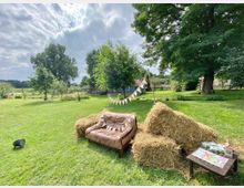 Eine weitläufige grüne Wiese mit großen Bäumen, einem braunen Ledersofa und Strohballen im Vordergrund, die mit einer Wimpelkette dekoriert sind. Im Hintergrund sind ein kleiner Holzzaun, ein Gebäude mit rotem Dach und eine ländliche Umgebung erkennbar.