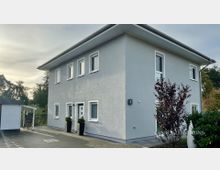 Zweistöckiges, modernes Wohnhaus mit hellgrauer Fassade und flachem Walmdach, mit mehreren rechteckigen Fenstern und einem Eingangsbereich mit Topfpflanzen. Davor liegt eine gepflasterte Einfahrt; links steht ein kleiner Carport/Schuppen, rechts sind Sträucher und Bäume im Gartenbereich zu sehen.